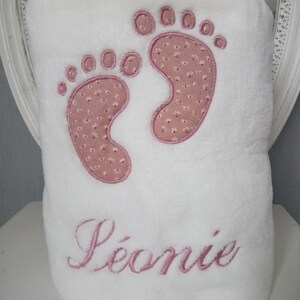 以下が含まれることがあります： ピンクの刺繍された足跡とピンクの「L&eacute;onie」という名前が書かれた白いベビーブランケットです。