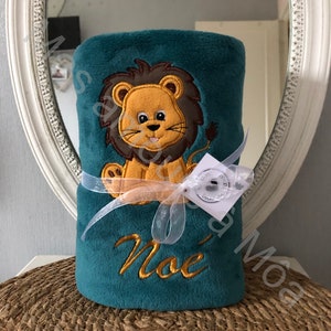 May include: A teal blue plush blanket with a lion applique and the name "Noe" embroidered on it. The blanket is tied with a white ribbon and a small tag.