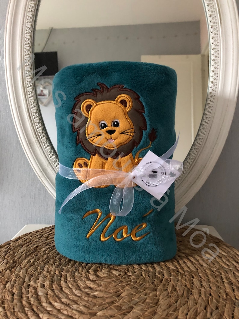 May include: A teal blue plush blanket with a lion applique and the name "Noe" embroidered on it. The blanket is tied with a white ribbon and a small tag.