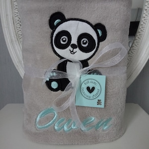 May include: A gray plush blanket with a black and white panda embroidered on it. The blanket is tied with a white ribbon and has the name "Owen" embroidered in light blue. A small tag with the text "Fait avec beaucoup d'amour" is attached to the ribbon.