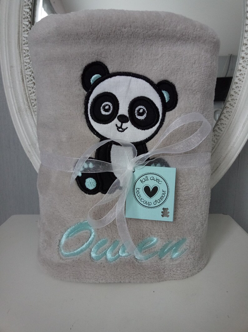 May include: A gray plush blanket with a black and white panda embroidered on it. The blanket is tied with a white ribbon and has the name "Owen" embroidered in light blue. A small tag with the text "Fait avec beaucoup d'amour" is attached to the ribbon.
