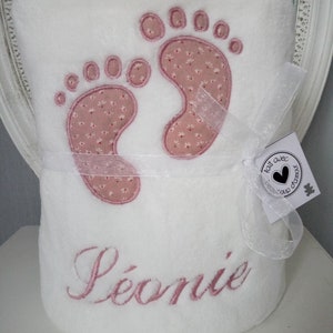 以下が含まれることがあります： ピンクの刺繍で足跡と「L&eacute;onie」という名前が刺繍された白いベビーブランケット。ブランケットは白いリボンで結ばれており、小さなタグにはフランス語で「fait avec beaucoup d'amour」と書かれています。