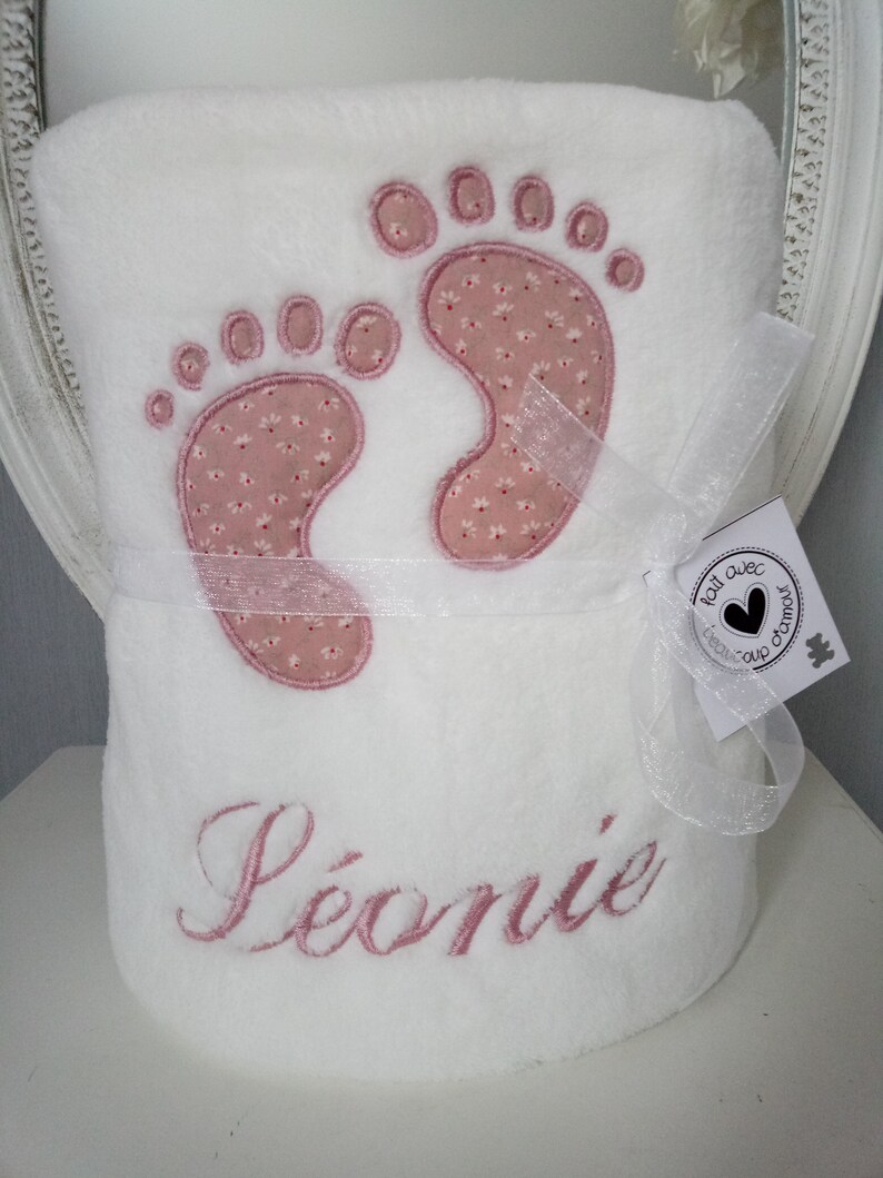 以下が含まれることがあります： ピンクの刺繍で足跡と「L&eacute;onie」という名前が刺繍された白いベビーブランケット。ブランケットは白いリボンで結ばれており、小さなタグにはフランス語で「fait avec beaucoup d'amour」と書かれています。