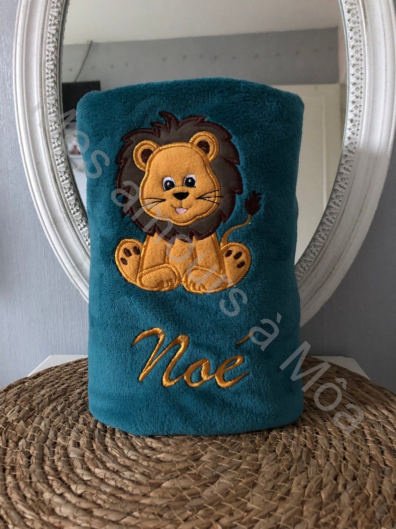May include: A teal blue plush blanket with an embroidered lion and the name "Noe" in gold thread.