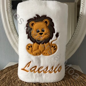 May include: White plush blanket with an embroidered lion in brown, yellow, and orange. The lion is sitting with its paws crossed and has a happy expression. The blanket also has the name "Laesso" embroidered in gold thread.