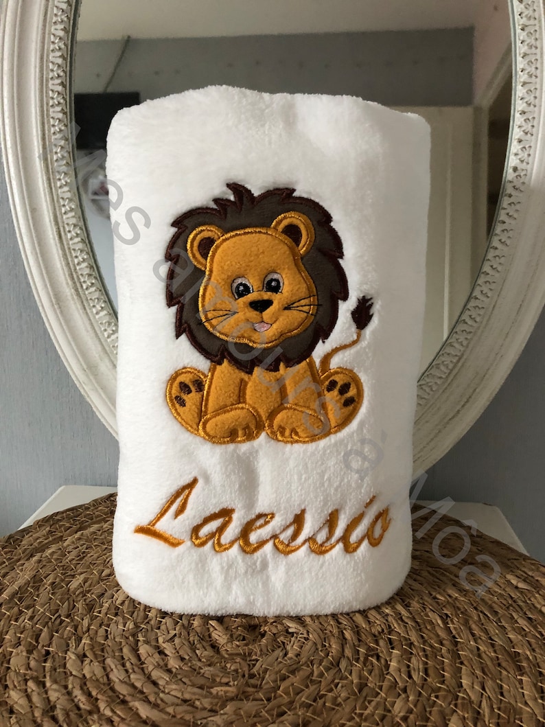 May include: White plush blanket with an embroidered lion in brown, yellow, and orange. The lion is sitting with its paws crossed and has a happy expression. The blanket also has the name "Laesso" embroidered in gold thread.