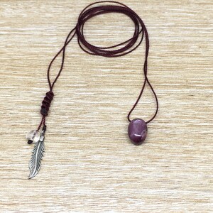 May include: A burgundy cord necklace with a purple gemstone pendant and a silver feather charm. The cord has a knot at the top and a clear crystal bead.
