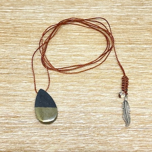Peut inclure: Un collier en cordon marron avec un pendentif en forme de larme en pierre noire et dorée. Le collier a un charme en plume argentée à l'extrémité.