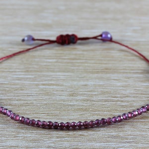 May include: A delicate bracelet made with small, faceted garnet beads strung on a red cord. The bracelet has a sliding knot closure with amethyst beads at each end.