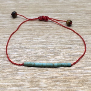 Op de afbeelding: Een rood koord armband met een turquoise kralenbalk en bruine kralen aan de uiteinden.