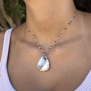 Peut inclure: Un collier avec un pendentif en forme de goutte d'eau et un fil d'argent. Le pendentif est blanc avec des veines grises. La chaîne du collier est une fine corde de couleur claire avec de petites perles noires et blanches.