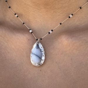 Peut inclure: Un collier artisanal avec un pendentif en forme de goutte d'eau présentant un motif blanc et gris, encadré dans une monture en fil de fer. Le collier a un cordon beige délicat avec des perles noires et blanches, créant une esthétique élégante et naturelle.
