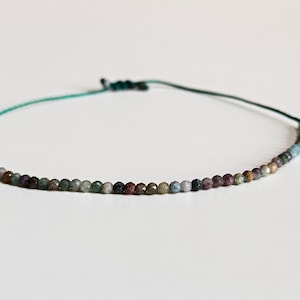Op de afbeelding: Een delicate kralenarmband met kleine, veelkleurige edelstenen geregen op een teal koord. De kralen vertonen tinten groen, bruin en paars. De armband is met een simpele knoop vastgemaakt.