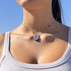 Può includere: Una delicata collana con un ciondolo a forma di goccia con un design blu e argento. Il ciondolo è avvolto in filo metallico e pende da una sottile catena d'oro con piccole perline nere. È visibile anche un orecchino abbinato con un piccolo ciondolo.