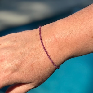 Peut inclure: Un bracelet délicat composé de petites perles magenta. Les perles sont enfilées ensemble, créant un accessoire subtil. Le bracelet est présenté sur un poignet, sur fond bleu.