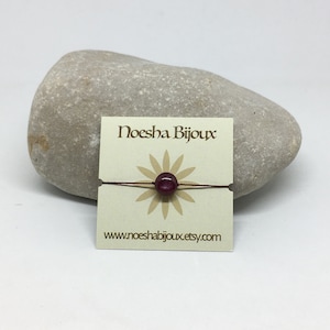 May include: A red string bracelet with a dark red bead and a white card with the text "Noesha Bijoux" and "www.noeshabijoux.etsy.com".