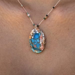Puede incluir: Un collar artesanal con un colgante ovalado con un mosaico de piedras azules, naranjas y blancas. El colgante está encerrado en una jaula de alambre y ensartado en un cordón color crema con pequeñas cuentas de colores.