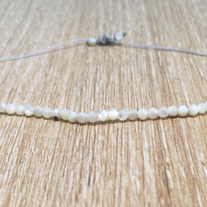 Puede incluir: Un collar blanco hecho de pequeñas cuentas redondas iridiscentes enhebradas en un cordón delgado gris.