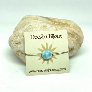 Puede incluir: Un brazalete de piedra azul claro con un cordón marrón. El brazalete está sobre una tarjeta con el texto "Noesha Bijoux" y "www.noeshabijoux.etsy.com".