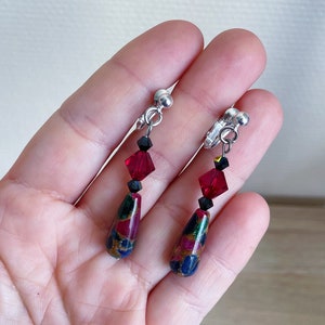 May include: A pair of clip-on earrings with a teardrop-shaped stone featuring a red, blue, and gold pattern. The earrings are accented with black and red crystal beads.