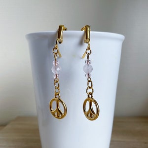 Könnte beinhalten: Ein Paar goldene Peace-Zeichen-Ohrringe mit rosa Perlen, die unten hängen.