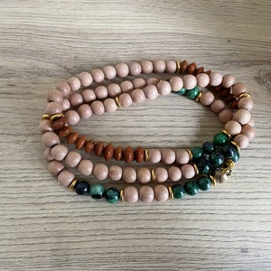 Puede incluir: Un brazalete de cuentas de madera de múltiples hilos con cuentas verdes y marrones y un dije dorado.