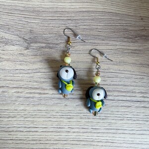 May include: A pair of dangle earrings featuring a small gray dog charm with a yellow tag. The earrings are made with silver-toned metal and have a light yellow bead accent.