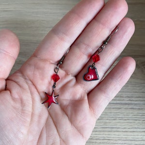 May include: A pair of red enamel earrings with a star and heart design. The star earring has a red crystal bead and the heart earring has a red crystal bead and a black eye detail.
