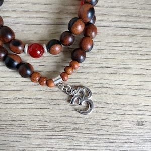 May include: A wooden mala necklace with brown and black beads, a red stone bead, and a silver Om charm.