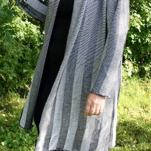 Puede incluir: Una persona con una camiseta negra y pantalones cortos negros, con una rebeca larga a rayas blancas y negras. Está de pie sobre hierba verde, con árboles y follaje al fondo. Lleva zapatos negros.