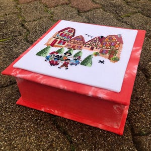 Peut inclure: Une boîte rouge avec un dessus brodé représentant un village avec des maisons, des arbres et des personnages. Les côtés de la boîte ont un effet tie-dye, et le dessus est une pièce séparée.