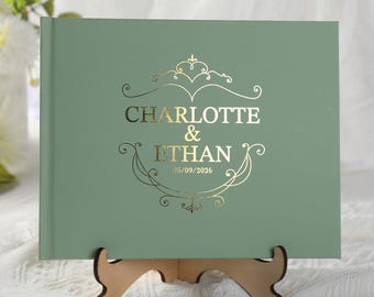 Può includere: Un libro degli ospiti rettangolare color verde salvia con scritte in lamina d'oro che riportano CHARLOTTE & ETHAN e la data 05/09/2026, circondato da una cornice in filigrana dorata, appoggiato su un piccolo supporto in legno.