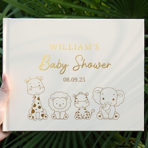 Puede incluir: Un libro de visitas rectangular blanco con texto en lámina dorada que dice WILLIAM'S Baby Shower 08.09.25. Debajo del texto hay ilustraciones de líneas doradas de una jirafa, un león, una cebra y un elefante sentados, con pequeños corazones.