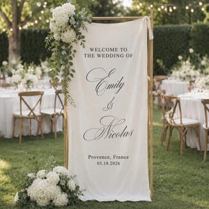 Puede incluir: Un cartel de bienvenida de boda vertical de tela color hueso colgado en un marco de madera, con texto negro que dice WELCOME TO THE WEDDING OF Emily & Nicolas, Provence, France, 18.05.2026, decorado con hortensias blancas y follaje verde.