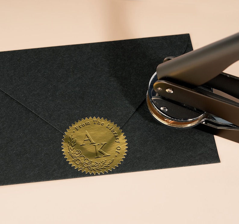 May include: A gold foil sticker with the text "From the Library of" and the initials "A.K." in a wreath design. The sticker is being applied to a black envelope with a handheld stamp.