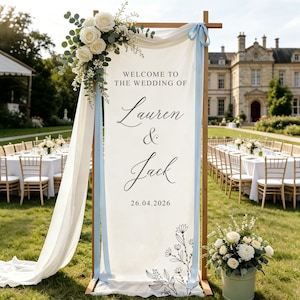 Op de afbeelding: Een verticaal welkomstbord voor een bruiloft op een houten frame met witte stof en de tekst "WELCOME TO THE WEDDING OF Lauren & Jack 26.04.2026". Het bord is versierd met witte rozen, eucalyptus en lichtblauwe linten.