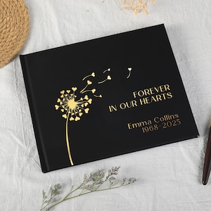 Könnte beinhalten: Ein schwarzes, rechteckiges Gedenkbuch mit goldfarbenem Textaufdruck FOREVER IN OUR HEARTS, Emma Collins, 1968-2025. Auf der linken Seite des Einbands befindet sich eine goldene Pusteblumen-Grafik mit herzförmigen Samen im Wind.