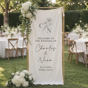 Op de afbeelding: Een verticaal trouwbord van gebroken wit textiel aan een houten frame, met zwarte tekst "WELCOME TO THE WEDDING OF Charles & Nina", de datum "03.12.2026", "Rome, Lazio" en een florale lijntekening met de initialen C en N.