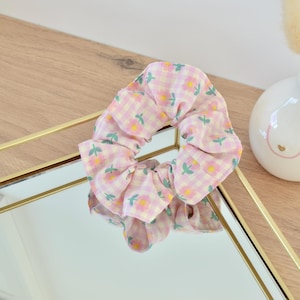 Könnte beinhalten: Ein rosa Vichy-Karo-Haargummi mit einem Blumenmuster aus gelben und grünen Blüten. Das Haaraccessoire ist aus weichem Stoff gefertigt und soll das Haar an Ort und Stelle halten. Das Haargummi wird auf einer spiegelnden Oberfläche gezeigt.