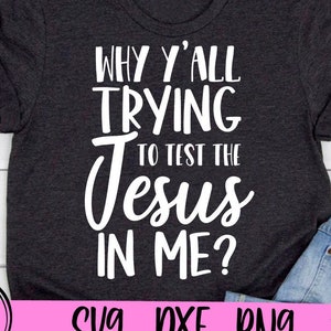 May include: A dark gray t-shirt with white text that reads "Why y'all trying to test the Jesus in me?" The shirt is on a wooden surface with a pair of blue jeans and a pink flower.