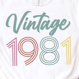 May include: White t-shirt with the words "Vintage 1981" in a retro design. The word "Vintage" is in a teal script font, and "1981" is in a multi-colored, striped font. A casual, everyday tee.