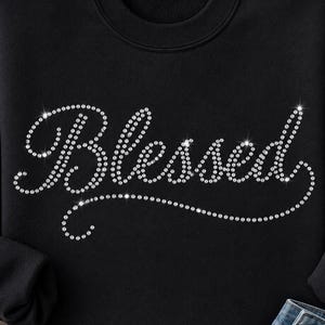 Könnte beinhalten: Ein schwarzes Langarm-Sweatshirt mit dem Wort Blessed in einer geschwungenen Schreibschrift. Der Schriftzug besteht aus kleinen, klaren, reflektierenden Strasssteinen, die die Buchstaben und einen dekorativen Unterstrich auf der Brust bilden.