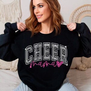 Op de afbeelding: Zwart sweatshirt met de woorden "Cheer Mama" in zilveren glitter en een roze glitterhart.