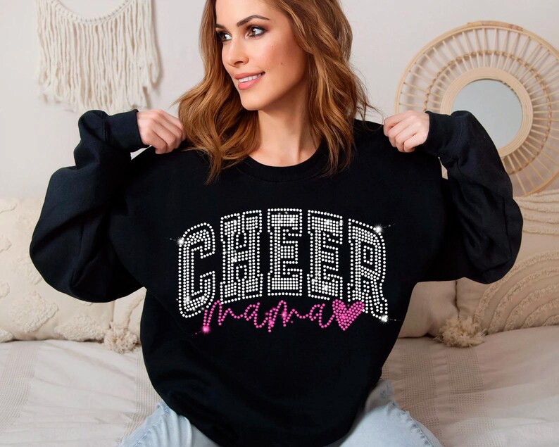 Op de afbeelding: Zwart sweatshirt met de woorden "Cheer Mama" in zilveren glitter en een roze glitterhart.