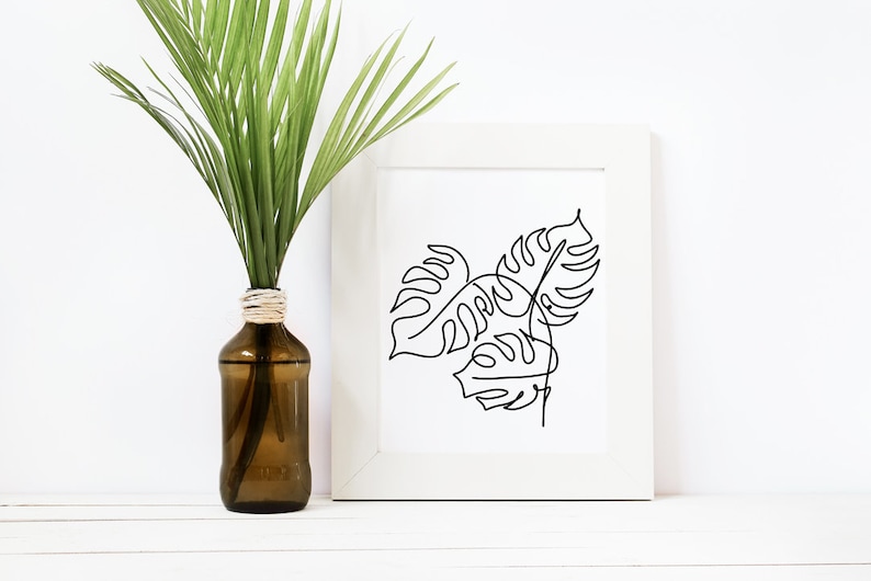 May include: A white frame with a black line drawing of three monstera leaves. The frame is sitting on a white surface next to a brown glass bottle with a green plant inside.