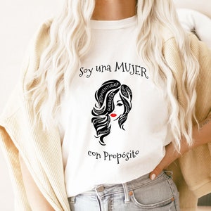 以下が含まれることがあります： 白いTシャツに、黒と白のグラフィックで女性の顔が描かれており、髪の周りに「Soy una MUJER con Prop&oacute;sito」というテキストが書かれています。