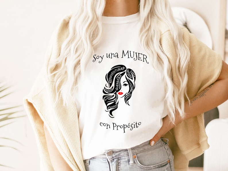 以下が含まれることがあります： 白いTシャツに、黒と白のグラフィックで女性の顔が描かれており、髪の周りに「Soy una MUJER con Prop&oacute;sito」というテキストが書かれています。