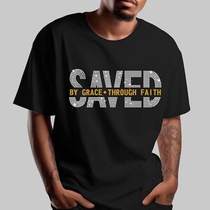 K&ouml;nnte beinhalten: Schwarzes T-Shirt mit dem Text "SAVED BY GRACE THROUGH FAITH" in Wei&szlig; und Gold. Der Text ist gestapelt, mit "SAVED" oben und unten. Das T-Shirt hat einen Rundhalsausschnitt.