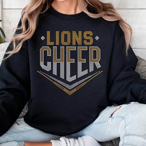 May include: Black sweatshirt with the words "LIONS CHEER" in gold and silver rhinestone-like lettering. The design includes a gold chevron at the bottom. The sweatshirt is a crew neck style.