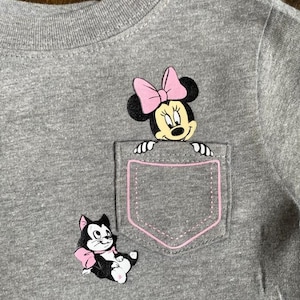 Puede incluir: Una camiseta gris con un bolsillo que presenta a una Minnie Mouse de dibujos animados asom&aacute;ndose por la parte superior y un gato de dibujos animados con un lazo rosa en la parte inferior.